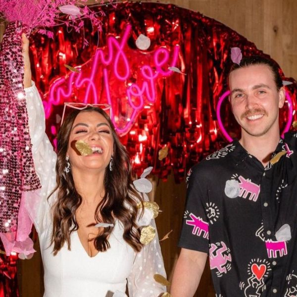 Crazy In Love Custom Neon® wedding sign in pink, styled against a red streamer background with the bride and groom in the foreground.