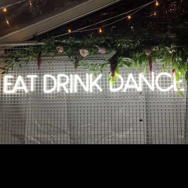 EAT DRINK DANCE large LED neon sign for events & weddings, shown on wire mesh with floral arrangement & fairy lights - photo from CustomNeon.com.au