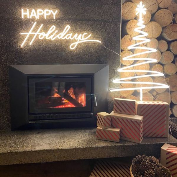 White LED neon Christmas tree & Happy Holidays sign mounted above a fireplace with wooden logs, presents & a basket of pinecones- by CUSTOM NEON® 