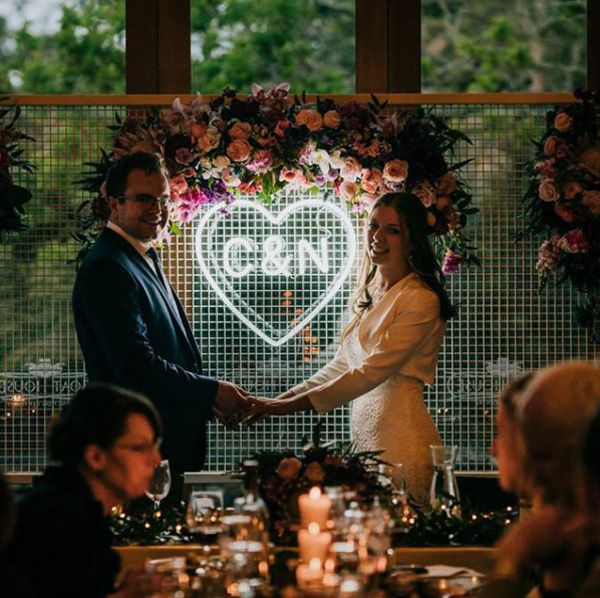Beautiful LED neon heart light personalised with the bride & groom's initials. Shown here on a mesh background surrounded by a floral arrangement. Photo from CustomNeon.com.au