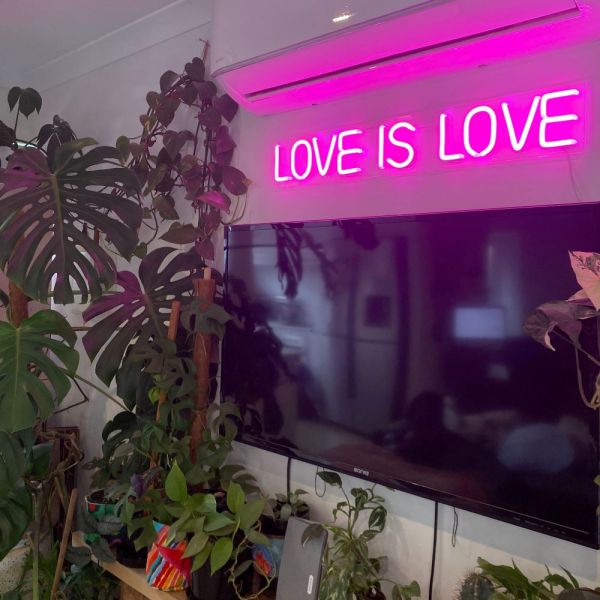Custom Neon® pink LOVE IS LOVE light sign shown wall mounted above a TV in a living room