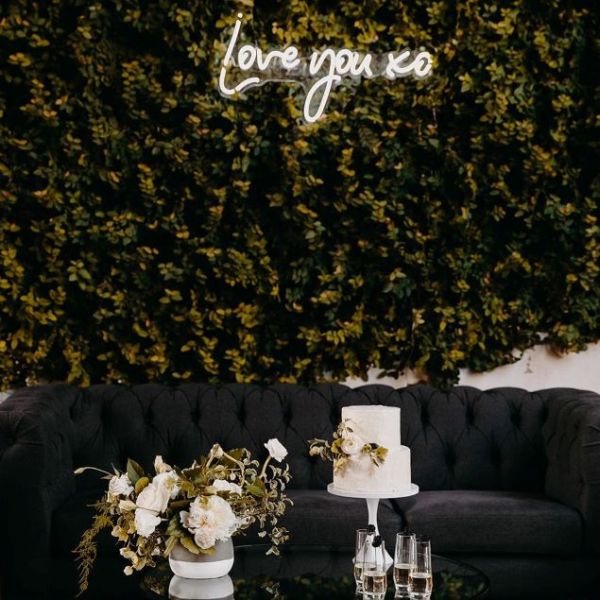 Love You XO LED neon sign on a green photo wall with a floral arrangement and cake in the foreground  - sign by CustomNeon.com.au / photo by candileonardphotography.com