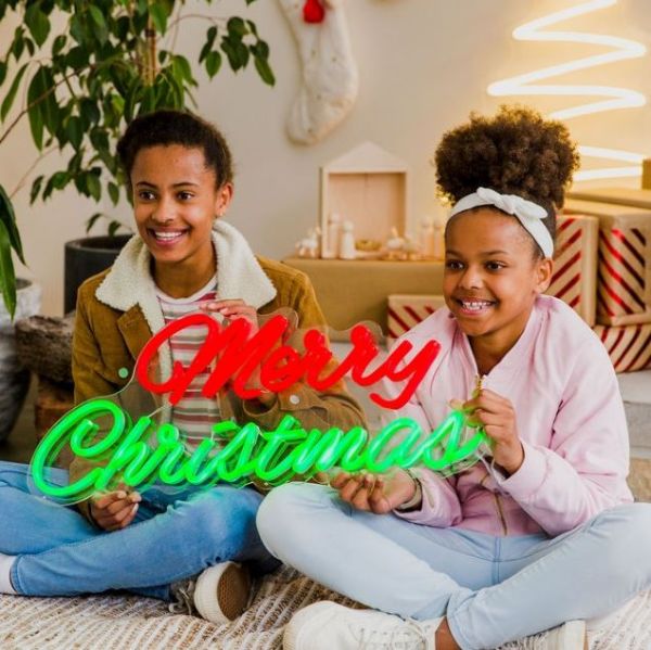 Merry Christmas Red & Green LED Neon Sign by CUSTOM NEON® held by smiling girls with a Nativity scene, presents & decorations in the background.
