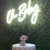 Oh Baby white LED neon sign shown on a green wall above a baby shower cake @CustomNeon