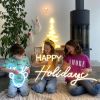 CUSTOM NEON® Holiday Lights: Reindeer Face, Happy Holidays & Modern LED Neon Xmas Tree held by children in front of a Nativity scene, stockings & wood burning stove