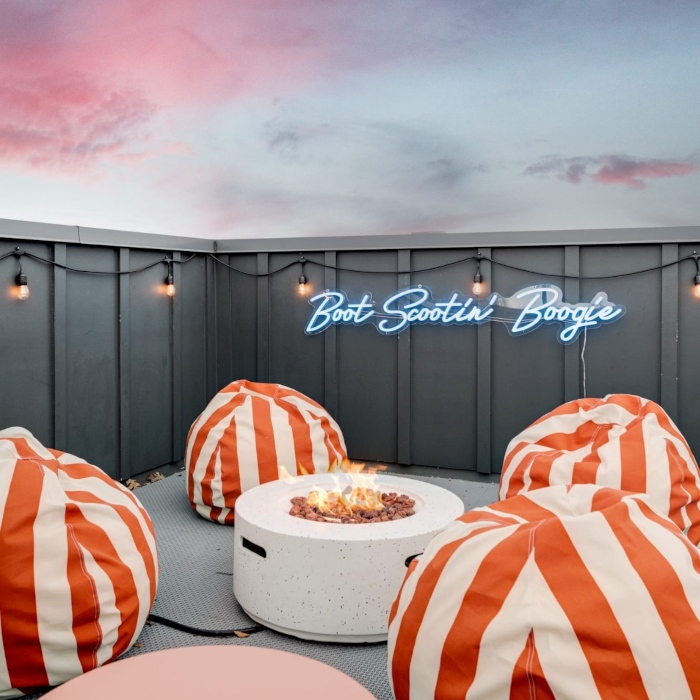 Boot Scootin Boogie light blue outdoor Custom Neon® sign on a balcony with beanbags, a firepit and a pink sunset overhead @Somerled