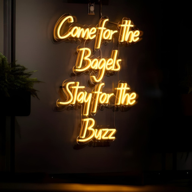 Yellow Custom Neon® sign on a black wall featuring the @buzzbagelz tagline