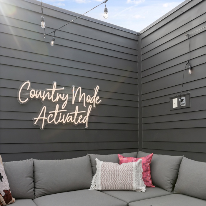 Country Mode Activated warm white weatherproof Custom Neon® sign on a grey panel fence with festoon lighting overhead in an outdoor space designed by Somerled