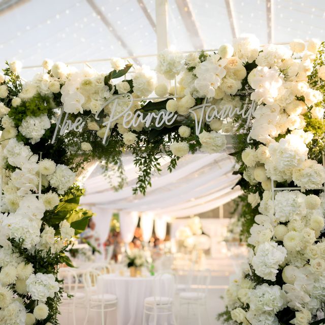 Family name sign in white by Custom Neon® attached to a frame at the entrance to the wedding marquee surrounded by white roses and florals @kaylaitsines