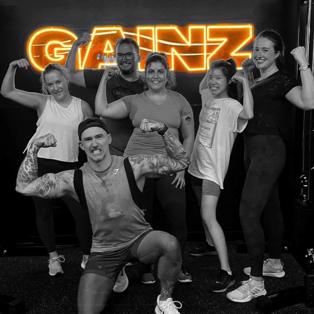 Custom Neon® dark yellow GAINZ sign on black wall behind gym goers @bodiesbyphil613