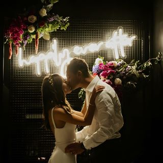 Custom Neon® warm white Happily Ever After quote sign on wire mesh surrounded by roses behind the newlyweds