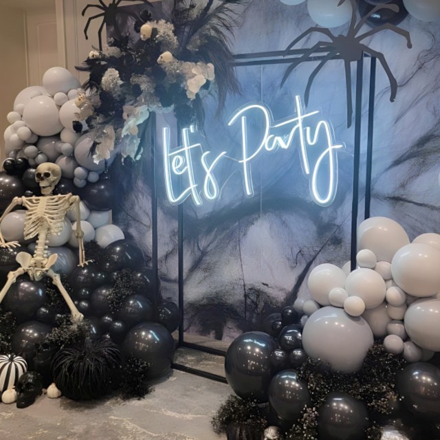 Let's Party Custom Neon® sign on a black frame surrounded by a black grey and white balloons and florals, skeletons and spiders props and styling by @decorandfiesta