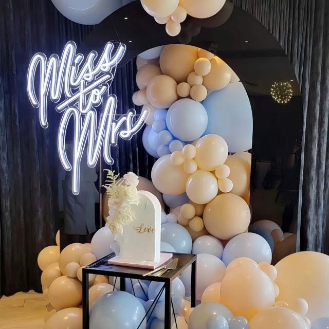 Custom Neon® white Miss to Mrs sign affixed to a reflective black arch surrounded by a balloon waterfall with a celebration cake in the foreground - event decor by @decorandfiesta