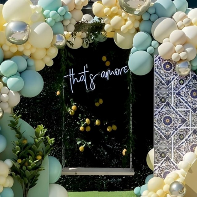 Custom Neon® white That's Amore sign on a green wall behind a wooden swing surrounded by a yellow and green balloon arch - event decor by @decorandfiesta