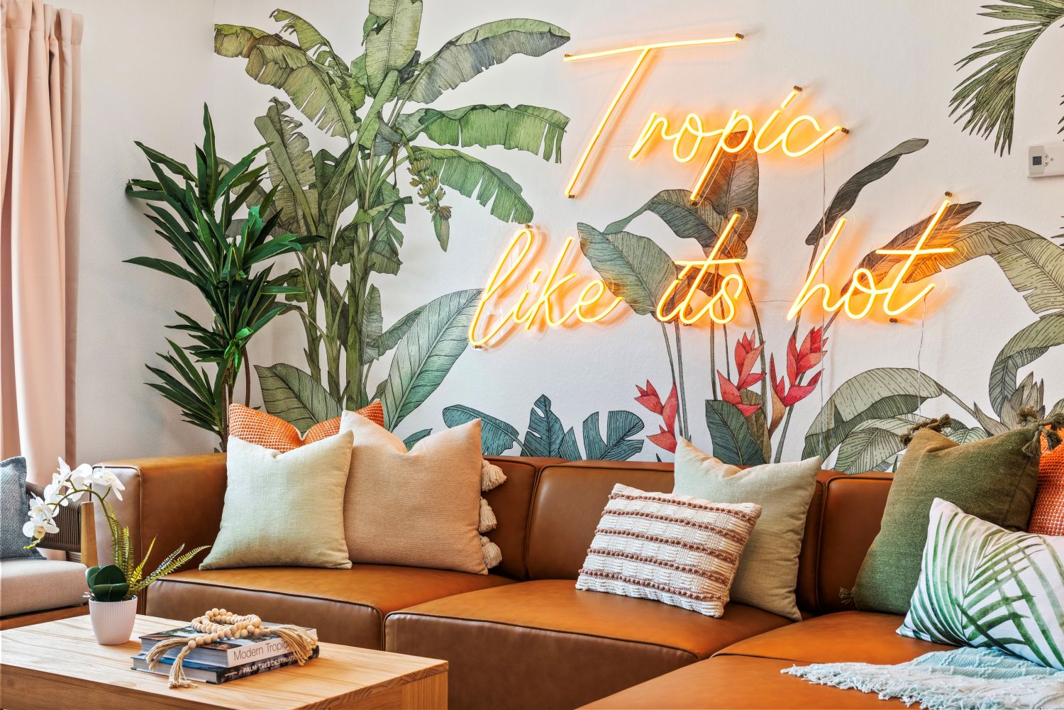 Tropic Like It's Hot orange LED Custom Neon® sign installed on a mural of tropical plants in a living room decorated with pink, green and tan colored furnishings designed by Somerled